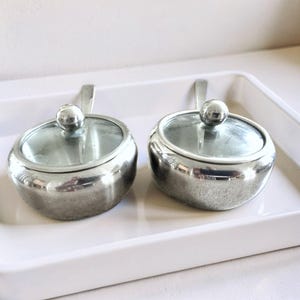 May include: Two round silver-toned metal condiment bowls with clear glass lids and small spherical knobs. Each bowl features a short, flat handle extending from the side, providing a polished, reflective surface for serving various food items.