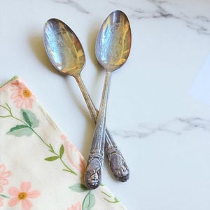 May include: Two vintage silver spoons with decorative handles and engraved details, crossed on a white marble surface. The spoons show signs of age and use. A floral patterned napkin in pink and green is in the lower left corner.