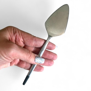 Może przedstawiać: Srebrna łopatka do ciasta z trójkątnym ostrzem i smukłym uchwytem. Uchwyt ma czarną końcówkę. Łopatka jest trzymana na białym tle.