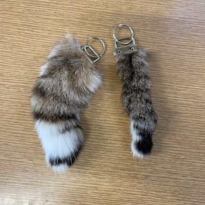 May include: Two brown and white faux fur keychains with silver key rings. The keychains are shaped like animal tails with black paw pads.