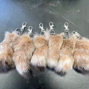 May include: A collection of seven keychains featuring small, furry animal paws. The paws are primarily light brown with darker tips and attached to silver-colored metal key rings with clasps. The keychains are arranged in a row.