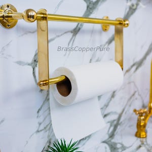 Puede incluir: Un portarrollos de papel de cocina dorado, montado en la pared, con un rollo de papel blanco. El soporte tiene una barra horizontal y soportes verticales. El fondo presenta una pared con vetas de mármol y un grifo dorado.