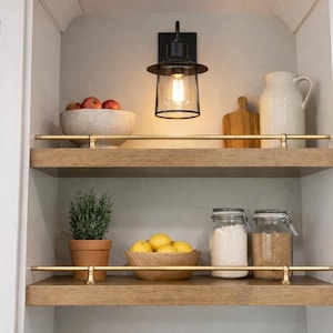 May include: A kitchen display with two wooden shelves featuring various items. The top shelf holds a bowl of apples, a cutting board, and a white pitcher. The bottom shelf displays a potted plant, a bowl of lemons, and glass jars.