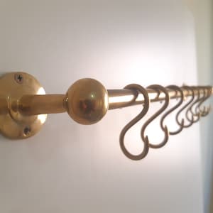May include: A gold metal wall-mounted hook rack with a round knob and multiple hooks. The rack is mounted on a white wall.