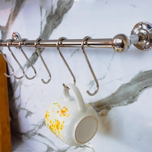 May include: A chrome kitchen rail with six S-hooks, mounted on a marble-effect wall. A white mug with yellow floral designs hangs from one of the hooks. A wooden chopping board is visible in the background.