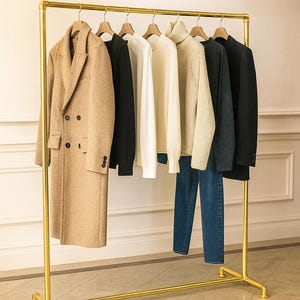 Pode incluir: Um cabide dourado com uma seleção de roupas. Um sobretudo bege, jaquetas pretas, suéteres brancos e creme e jeans azuis pendurados em cabides de madeira. O cabide está em um chão ladrilhado.