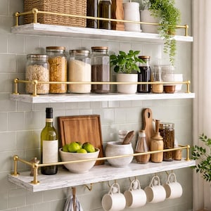 Puede incluir: Estanterías de cocina con superficies efecto mármol y barandillas doradas. Las estanterías contienen varios artículos de cocina, incluyendo tarros de granos, especias y plantas. También se exhiben una cesta tejida, botellas y un cuenco de fruta.