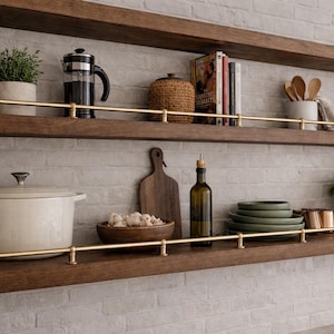 May include: Wooden kitchen shelves displaying various items. A French press, potted plant, and books are on the top shelf. The bottom shelf holds a Dutch oven, cutting board, and olive oil bottle. The shelves have brass rails.