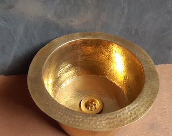 May include: A hammered brass sink with a round shape and a drain in the center. The sink has a gold finish and a textured surface.