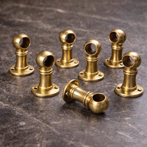 May include: A set of seven brass curtain rod brackets, each featuring a cylindrical base with a circular mounting plate and a spherical top section with a hollow center for holding a rod. The metal has a polished, reflective gold-toned finish.
