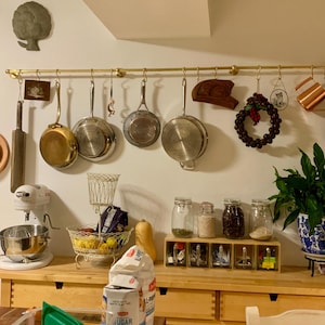 Op de afbeelding: Een keuken met een messing pottenrek dat boven een houten aanrecht hangt. Het rek bevat verschillende potten en pannen, waaronder een koperen pot, een roestvrijstalen pot en een gietijzeren koekenpan. Er staan ook verschillende potten met kruiden en een keukenmachine op het aanrecht.
