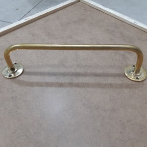 May include: A gold-toned metal towel bar with rounded ends. The bar is mounted on a light brown wall.