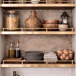 Puede incluir: Una estantería de cocina exhibe varios objetos. Hay un tarro de cristal con pasta, una jarra tejida, cuencos de madera y una linterna negra. Otros artículos incluyen botellas, platos, una cesta de huevos y un recipiente con la inscripción "PURE TEA".