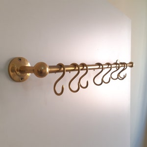May include: A brass kitchen utensil rack with a round knob and multiple hooks. The rack is mounted to a white wall.
