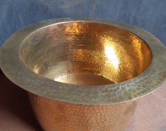 May include: A hammered brass bowl with a wide rim. The interior of the bowl is a bright gold color, reflecting light. The exterior has a textured, dimpled surface. The bowl is round and has a simple, elegant design.