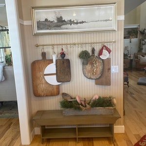 May include: A rustic wooden bench with a shelf underneath, decorated with a wooden box filled with greenery and candy canes. The wall behind the bench is adorned with a framed print, a row of hooks, and several wooden cutting boards.
