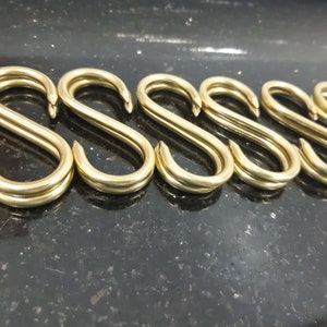 Może przedstawiać: Zbliżenie kilku mosiężnych haczyków w kształcie litery S ułożonych w rzędzie. Haczyki wykonane są z błyszczącego, metalicznego materiału i mają gładką, zakrzywioną konstrukcję. Tło to ciemna, nakrapiana powierzchnia.