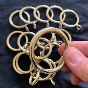 Peut inclure: Un ensemble d'anneaux de rideaux de couleur laiton. Les anneaux sont circulaires avec une petite boucle attachée. Un anneau est tenu dans une main, avec plusieurs autres en arrière-plan. Les anneaux ont une finition métallique brillante.