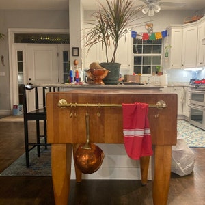 Puede incluir: Una isla de cocina de madera de carnicero con una barandilla y ganchos de latón. Una toalla de cocina a rayas rojas y blancas cuelga de la barandilla. Un cucharón de cobre cuelga de uno de los ganchos.