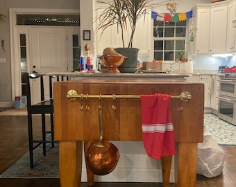May include: A wooden butcher block kitchen island with a brass rail and hooks. A red and white striped kitchen towel hangs on the rail. A copper ladle hangs from one of the hooks.