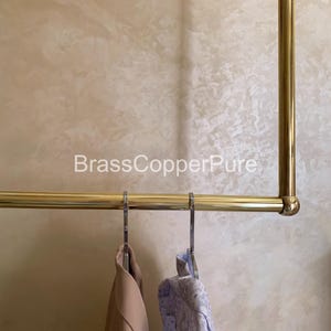以下が含まれることがあります： 真鍮製の洋服ラック。金属製のハンガーに2着の衣服が掛かっています。1つはライトブラウンのジャケット、もう1つは青と白の模様のシャツです。「BrassCopperPure」の文字が見えます。