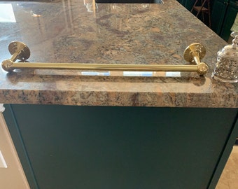 May include: A gold-toned towel bar with rounded ends is mounted on a granite worktop. The worktop is a dark brown with flecks of lighter brown and grey. The towel bar is mounted on a dark green cabinet.