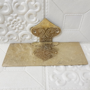 May include: A gold-colored metal wall mounted shelf with an ornate design. The shelf is rectangular with a decorative top piece in the shape of a pointed arch.
