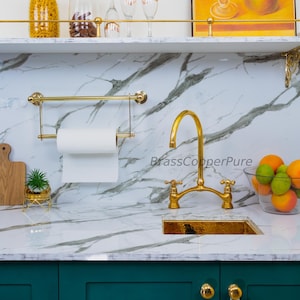 Pode incluir: Uma cena de cozinha com uma torneira e pia douradas, um porta-toalhas de papel e uma bancada de mármore. Uma pequena planta num vaso dourado e uma tigela de fruta também são visíveis. O texto "BrassCopperPure" está na bancada.