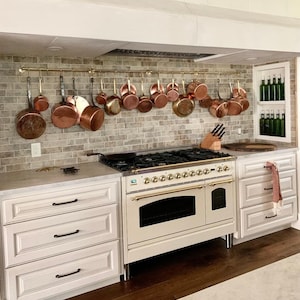 Könnte beinhalten: Eine weiße Küche mit einem cremefarbenen Herd mit goldenen Akzenten. Der Herd hat einen Doppelbackofen und ein Gaskochfeld. Über dem Herd hängt ein Gestell mit Kupfertöpfen und -pfannen an einer Messingstange.