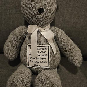 May include: A gray teddy bear made from knitted fabric, with black button eyes and a black stitched nose. It has a white ribbon around its neck and a patch with text. The text reads "sweater to wear...I'm there."