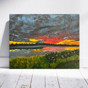 May include: Square canvas painting depicting a sunset over a lake. The sky features dark blue, grey, yellow, and red tones. The lake mirrors the sky's colours, with green foliage in the foreground. The artwork is displayed against a white wall.