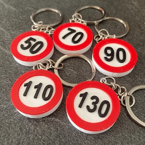 Peut inclure: Porte-clés avec des panneaux de limitation de vitesse. Chaque porte-clés présente un cercle blanc avec un chiffre noir à l'intérieur d'un cercle rouge. Les chiffres sont 50, 70, 90, 110 et 130. Chaque porte-clés a un anneau argenté.