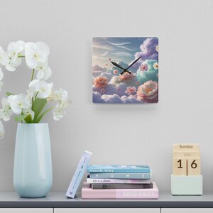 May include: Square wall clock with a whimsical design featuring fluffy clouds, pastel flowers, and a blue sky. The clock hands are black. The clock is hanging on a white wall. The clock measures approximately 10 inches by 10 inches.