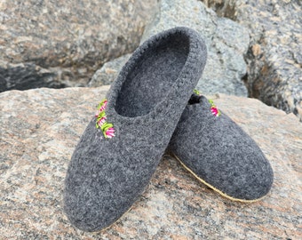 May include: A pair of gray felt slippers with floral embroidery in pink, green, and white. The slippers have a rounded toe and a light-colored sole. The background features large gray rocks.