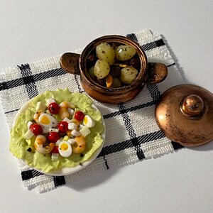 Op de afbeelding: Miniatuur poppenhuis voedselset met een salade met garnalen, tomaten en eieren op een bord, en een koperen pot met druiven. De set is geplaatst op een zwart-wit geruite servet. De miniatuur voedsel items zijn gedetailleerd.