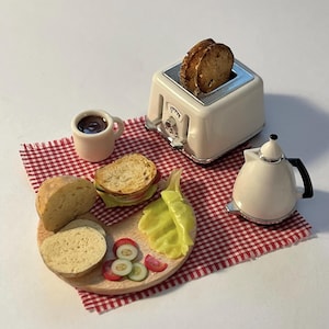 Op de afbeelding: Miniatuur ontbijt met een witte broodrooster met geroosterd brood, een kleine koffiekop, een theepot en een houten bord met brood, een sandwich en gesneden groenten. De scène staat op een rood-wit geruite doek.