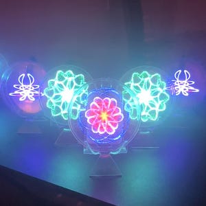 Puede incluir: Una colección de obras de arte circulares iluminadas. Cada pieza presenta un diseño central, con un patrón floral en el centro. Las piezas están iluminadas con colores neón vibrantes, incluyendo azul, verde y rosa.