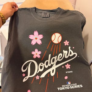 May include: Dark gray t-shirt featuring the word "Dodgers" in white script, a baseball, and pink cherry blossoms. The shirt also includes the text "MLB World Tour Tokyo Series".