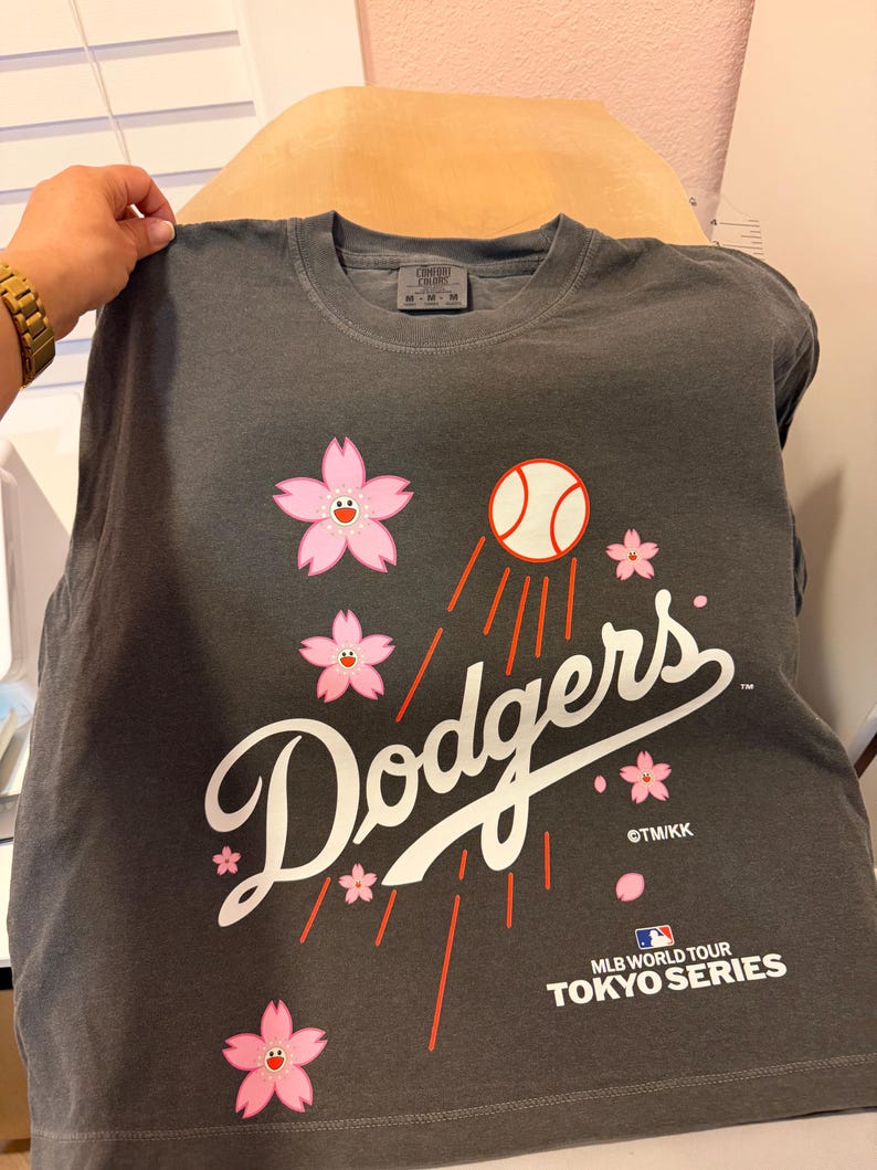 May include: Dark gray t-shirt featuring the word "Dodgers" in white script, a baseball, and pink cherry blossoms. The shirt also includes the text "MLB World Tour Tokyo Series".