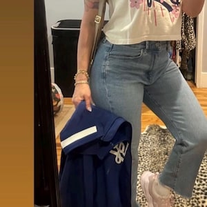 May include: A cream-colored crop top with the word "Dodgers" in blue script and pink floral accents. The person is wearing light blue jeans, pink sneakers, and holding a navy blue jacket.