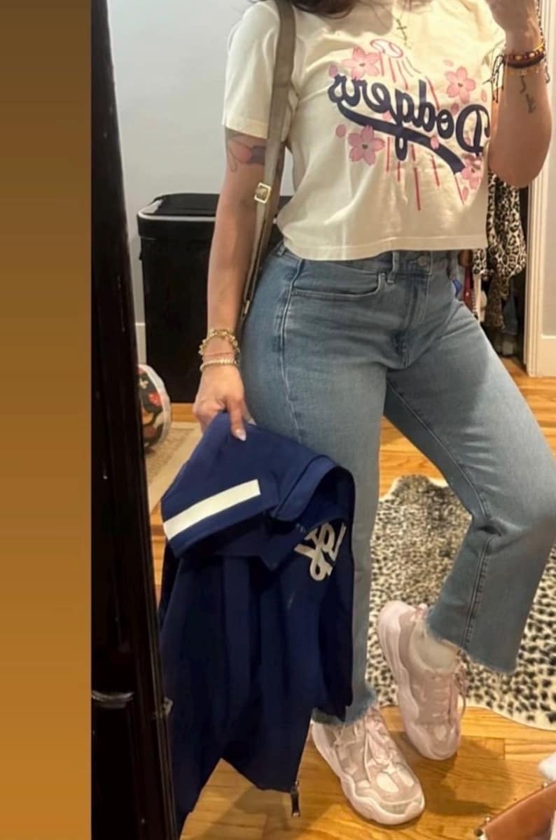 May include: A cream-colored crop top with the word "Dodgers" in blue script and pink floral accents. The person is wearing light blue jeans, pink sneakers, and holding a navy blue jacket.
