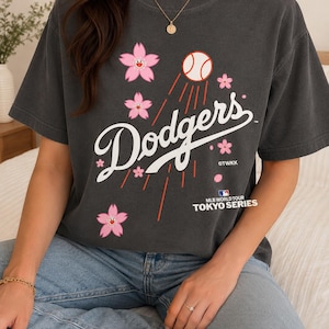 May include: Dark gray t-shirt featuring the word "Dodgers" in white script, with a baseball and pink cherry blossom graphics. The shirt also includes the text "MLB World Tour Tokyo Series".