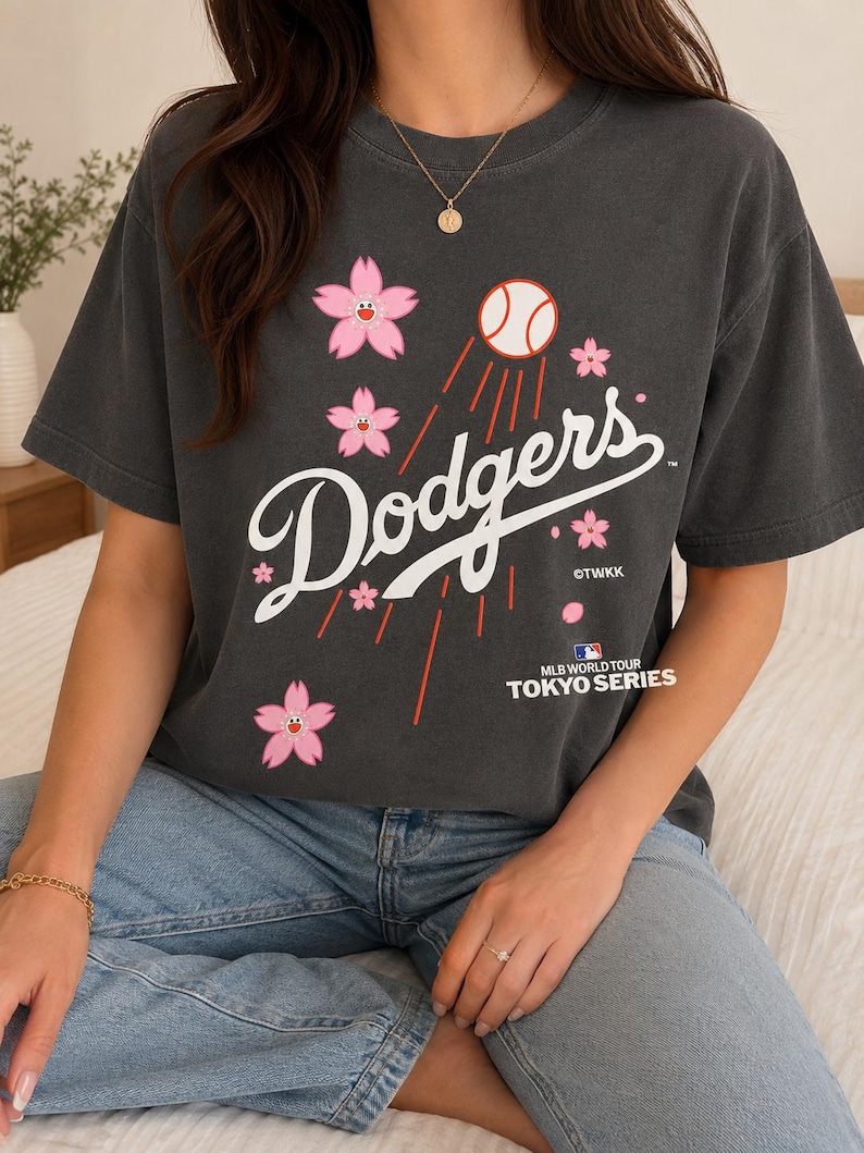 May include: Dark gray t-shirt featuring the word "Dodgers" in white script, with a baseball and pink cherry blossom graphics. The shirt also includes the text "MLB World Tour Tokyo Series".