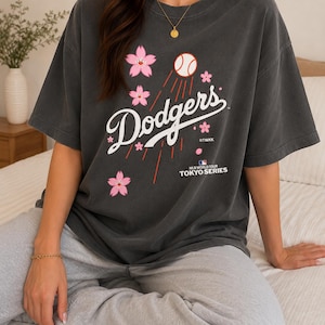 May include: Dark gray t-shirt featuring the word "Dodgers" in white script, with a baseball and pink cherry blossoms. The shirt also includes the text "MLB World Tour Tokyo Series".