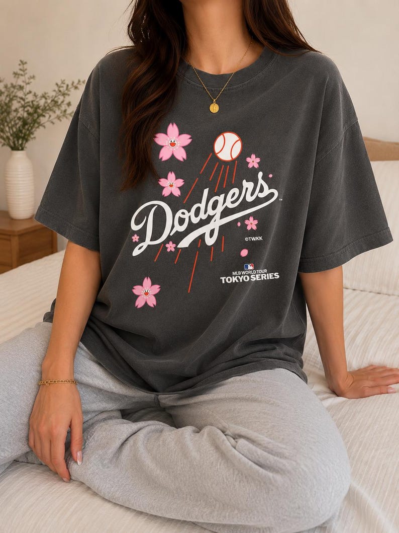 May include: Dark gray t-shirt featuring the word "Dodgers" in white script, with a baseball and pink cherry blossoms. The shirt also includes the text "MLB World Tour Tokyo Series".