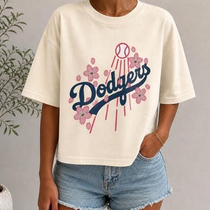 May include: Cream-colored t-shirt featuring the word "Dodgers" in navy blue script, with a baseball and pink cherry blossoms. The shirt has short sleeves and a relaxed fit, perfect for casual wear.