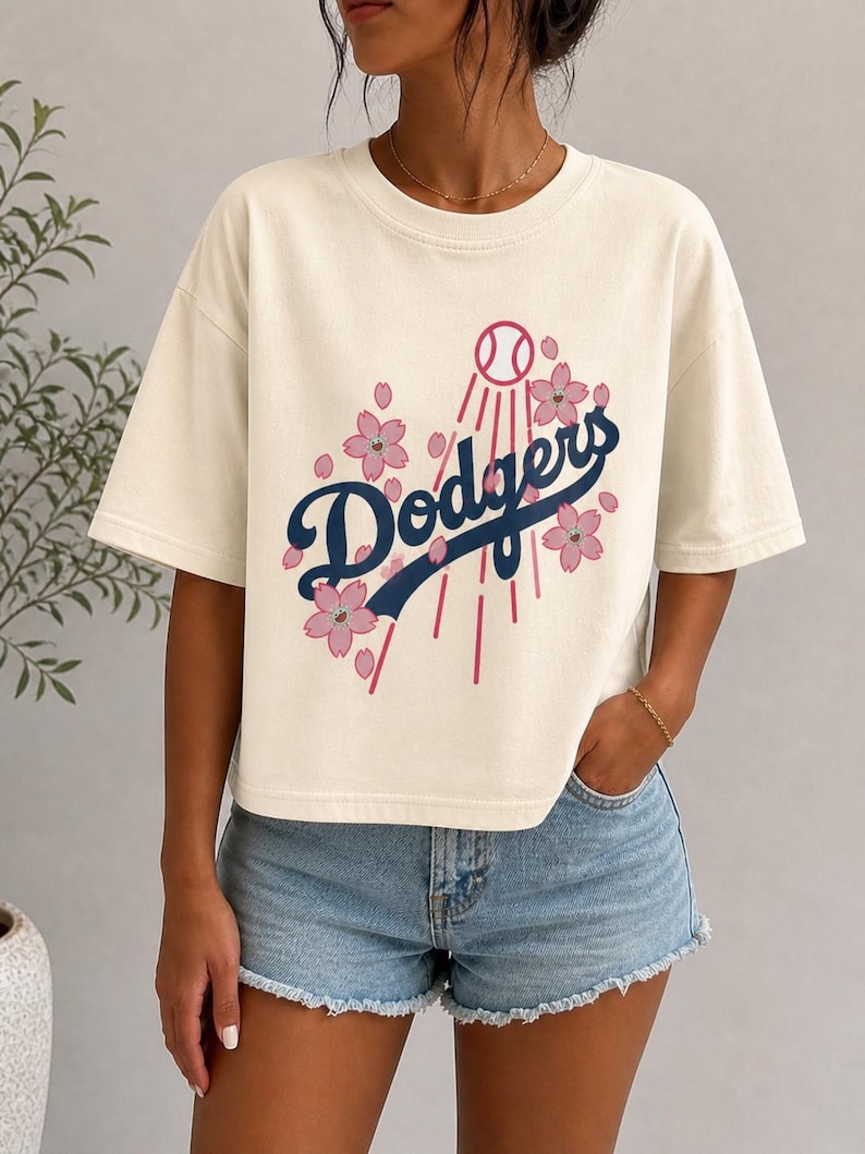 May include: Cream-colored t-shirt featuring the word "Dodgers" in navy blue script, with a baseball and pink cherry blossoms. The shirt has short sleeves and a relaxed fit, perfect for casual wear.
