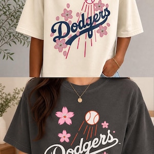 May include: Two short-sleeved t-shirts, one cream and one charcoal, featuring the word "Dodgers" in a script font. The design includes a baseball, pink cherry blossoms, and red lines. The charcoal shirt also has "MLB World Tour Tokyo Series" text.