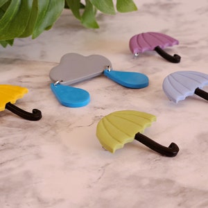 May include: Five colorful, handmade polymer clay pins. The pins are shaped like umbrellas and a cloud with raindrops. The umbrellas are in shades of purple, blue, yellow, and green. The cloud is gray with blue raindrops.
