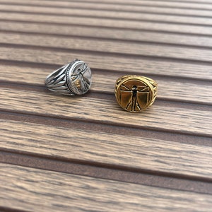 May include: Two silver and gold rings with a raised image of a man with outstretched arms. The rings are on a wooden surface.